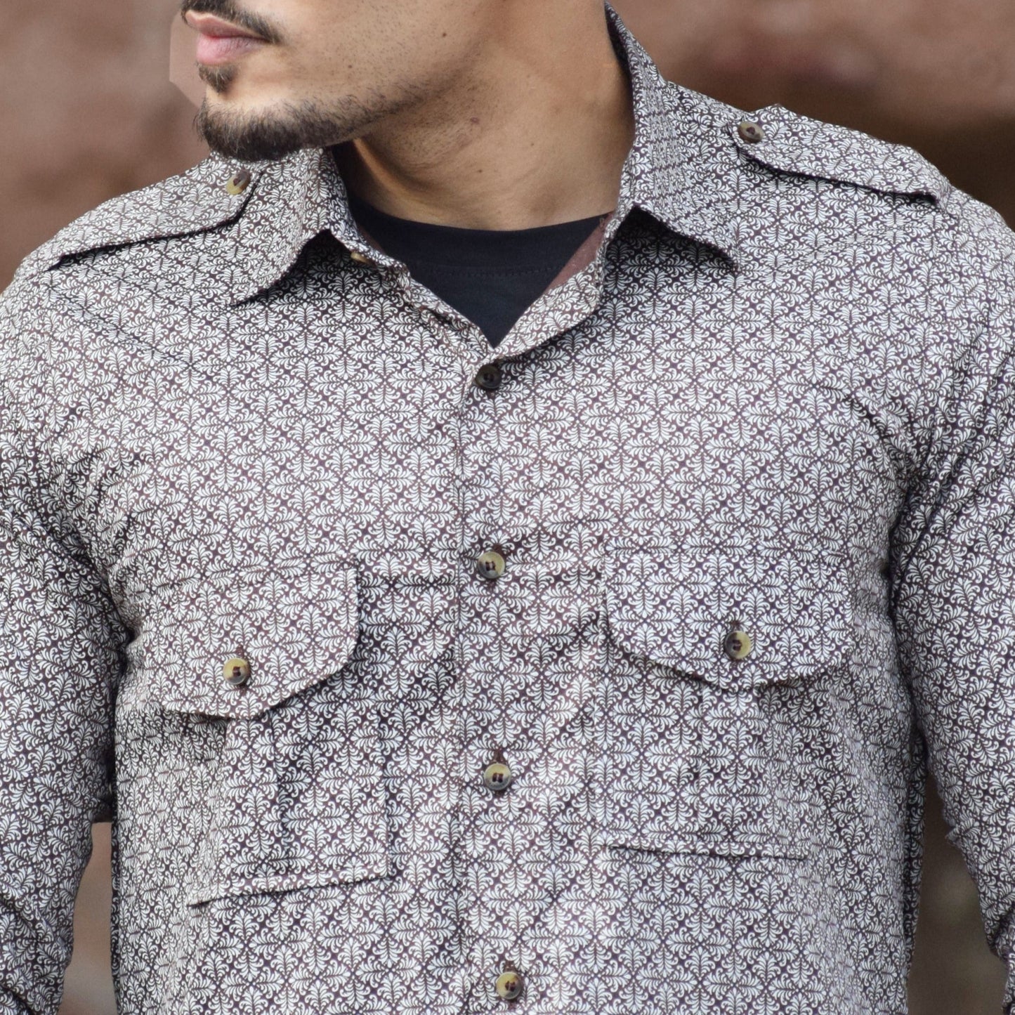 Man wearing a patterned shirt with rolled-up sleeves against a textured wall.