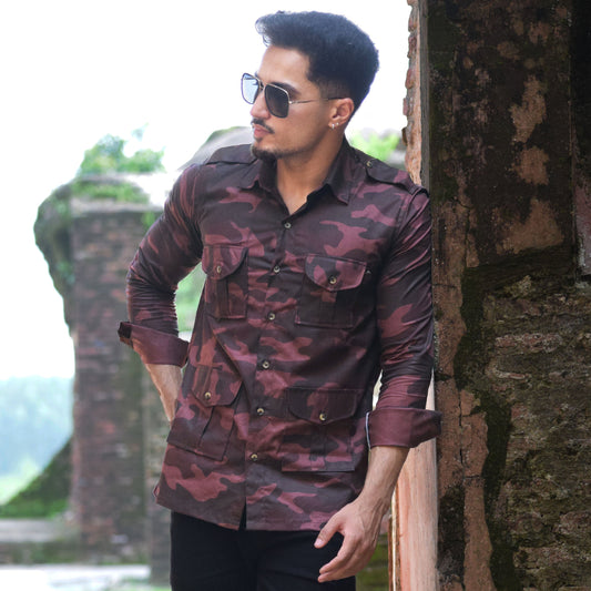 Man in a patterned shirt and black pants leaning against a stone wall with a scenic background.
