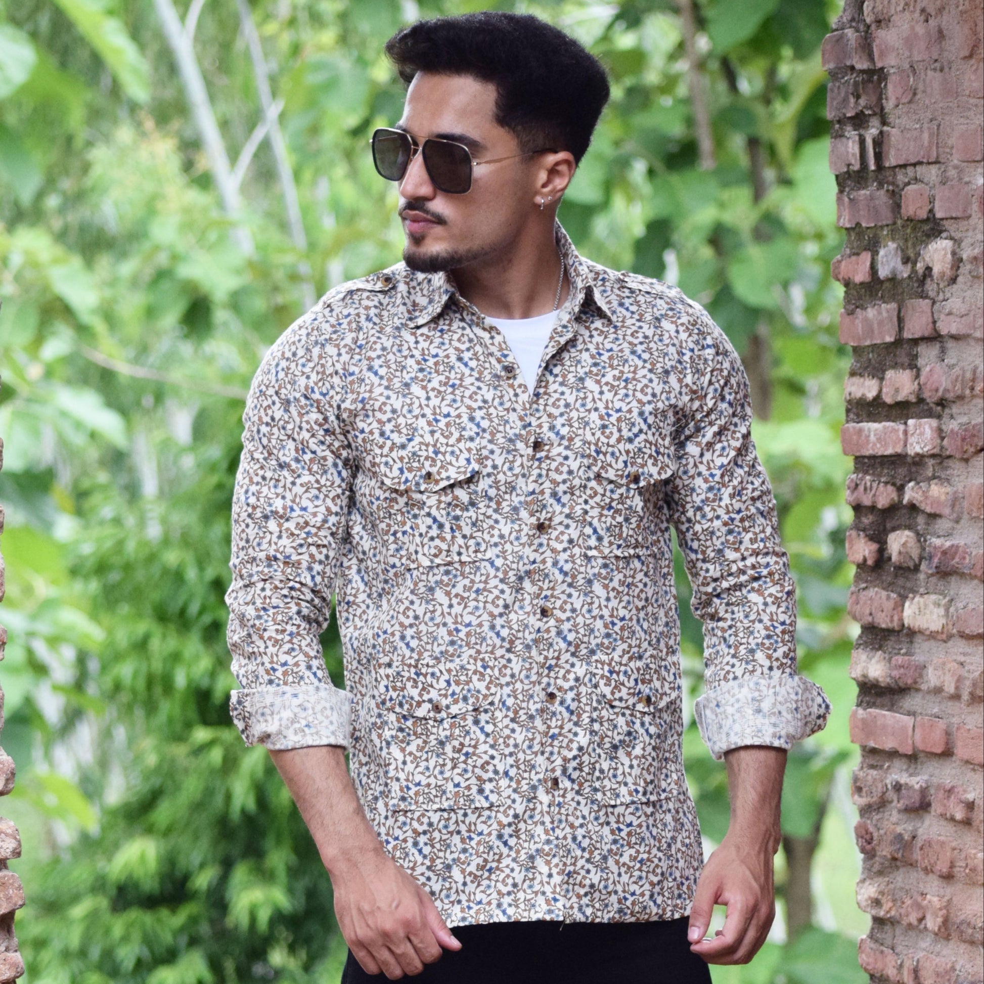 Man wearing a patterned shirt and sunglasses standing between two brick walls with greenery in the background