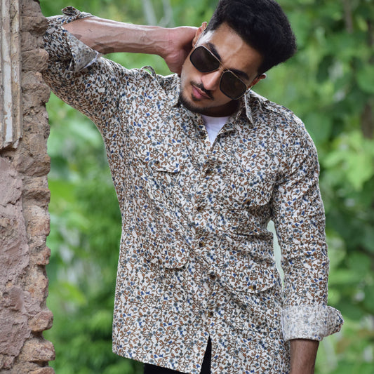Man wearing a patterned shirt and sunglasses, leaning against a stone wall with greenery in the background