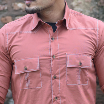 Man wearing a pink shirt with multiple pockets against a brick wall.