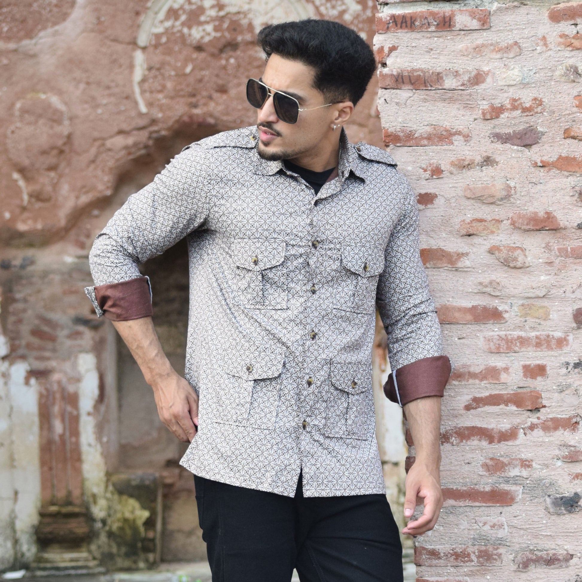 Man wearing a patterned shirt and sunglasses leaning against a textured wall.
