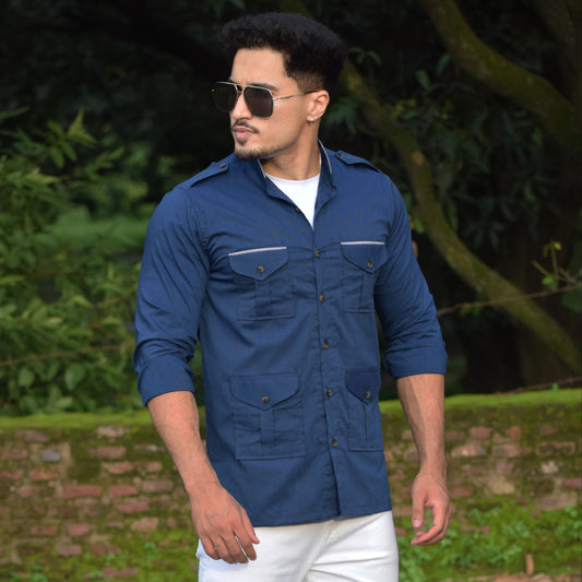Man wearing a blue shirt and white pants standing outdoors with greenery in the background