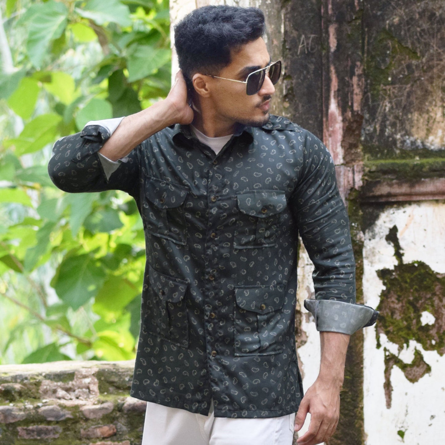 Man in a patterned shirt and white pants standing outdoors near a textured wall.