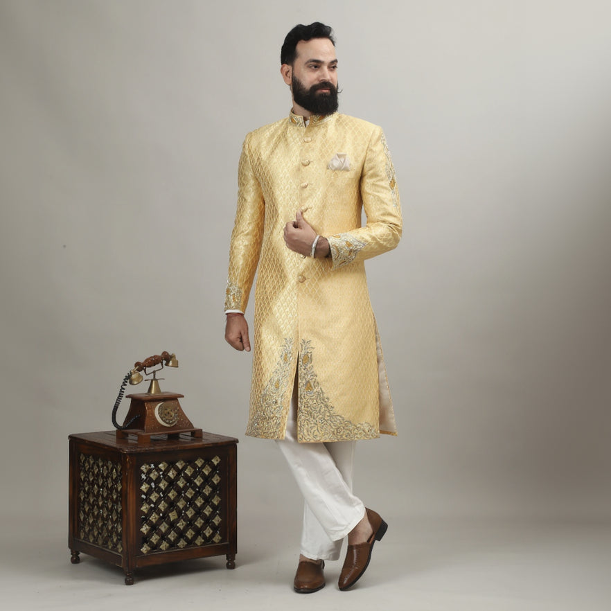 Man in a gold sherwani standing next to a wooden cabinet with intricate carvings on a plain background