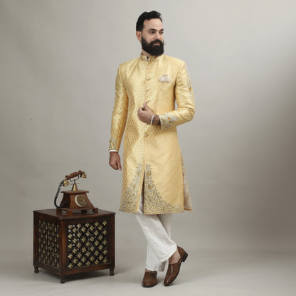 Man in a gold sherwani standing next to a wooden cabinet with intricate carvings on a plain background