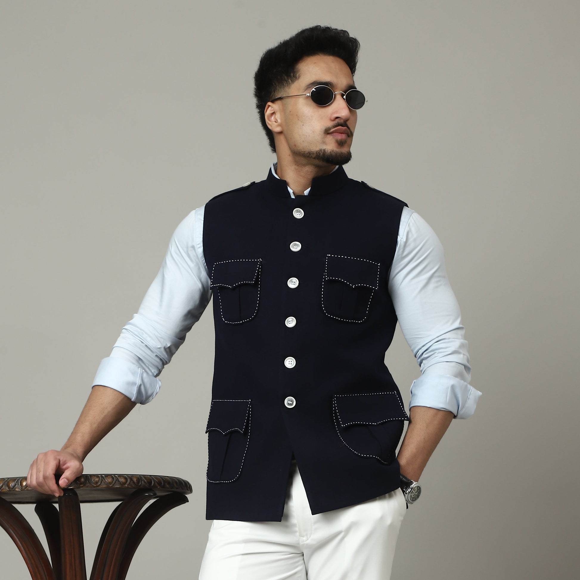 Person wearing a navy blue vest and white pants holding onto a wooden stool against a plain background