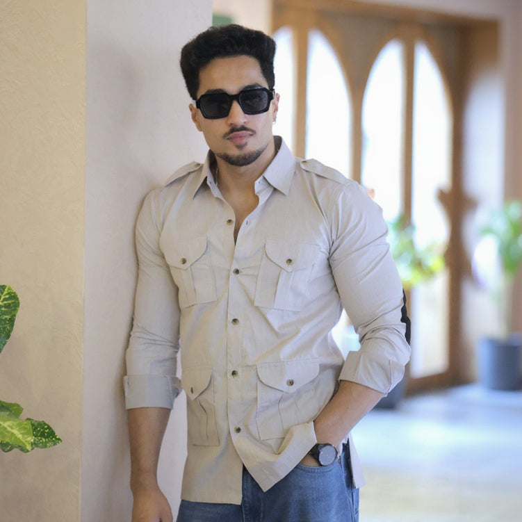 Man wearing sunglasses and a light-colored shirt leaning against a wall indoors.