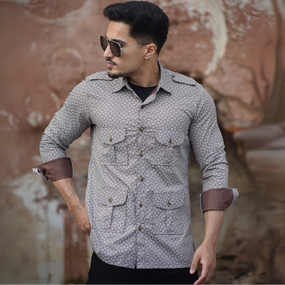 Man wearing a patterned shirt with rolled-up sleeves in front of a textured wall.