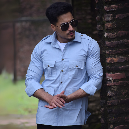 Man wearing sunglasses and a light blue shirt leaning against a brick wall.